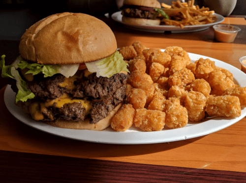 A large cheeseburger with lettuce and tomato, served with a side of crispy tater tots on a white plate.