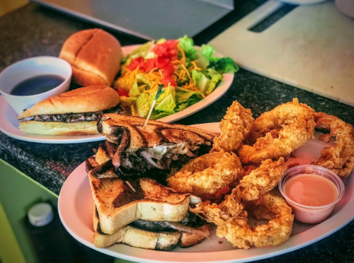 A plate with a grilled sandwich, onion rings, and a side of dipping sauce, alongside a salad and a sandwich.