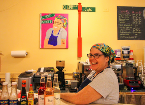 A smiling barista stands behind a counter with various drink bottles, next to a colorful portrait on the wall.