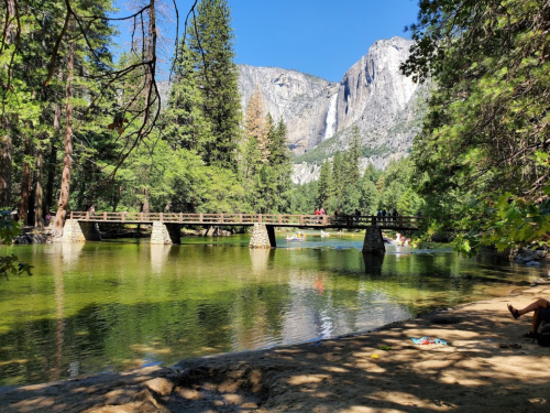 A serene river scene with a wooden bridge, surrounded by trees and mountains, with people enjoying the water.