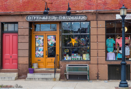 Colorful storefronts with a sign reading "Different Drummer," featuring vibrant decorations and a bench outside.