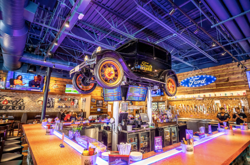 A vibrant bar interior featuring a vintage car suspended above the counter, with colorful lighting and screens in the background.