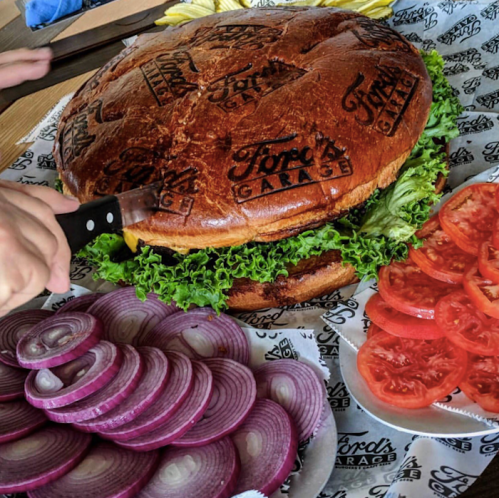 A large burger with a branded bun, surrounded by sliced onions, tomatoes, and lettuce on a patterned tablecloth.