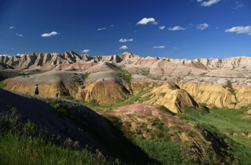 A person stands on a colorful hillside in a vast, rugged landscape under a blue sky with scattered clouds.
