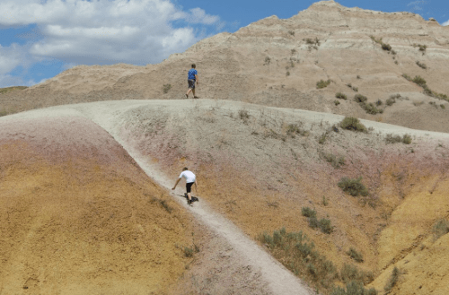 Two people hike on colorful, layered hills under a blue sky with clouds.