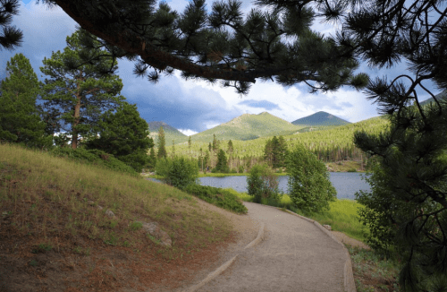 A scenic path by a lake, surrounded by lush greenery and mountains under a cloudy sky.