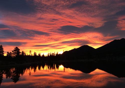 A vibrant sunset reflects on a calm lake, surrounded by silhouettes of trees and mountains.