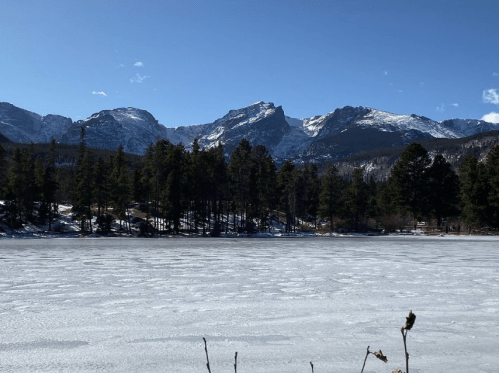 Snow-covered mountains rise above a frozen lake, surrounded by evergreen trees under a clear blue sky.