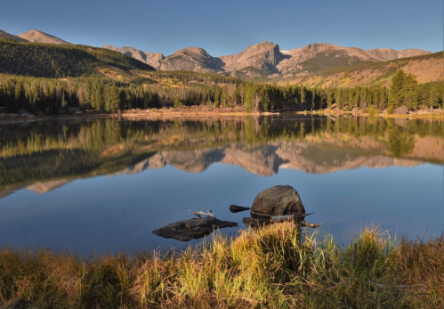 A serene lake reflects mountains and trees, with a rock in the foreground and autumn colors in the grass.