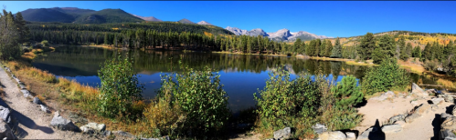 A serene lake surrounded by trees and mountains, reflecting autumn colors under a clear blue sky.