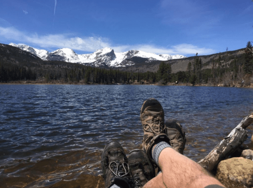 A person relaxes by a lake, feet in hiking boots resting on the shore, with snow-capped mountains in the background.