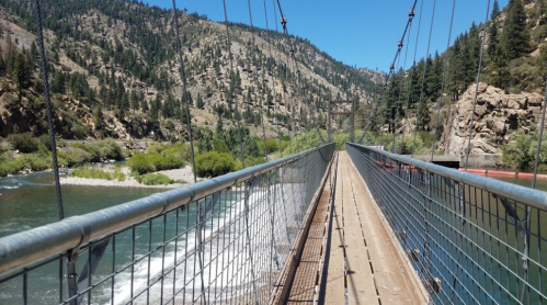 A suspension bridge spans a river, surrounded by lush greenery and mountains under a clear blue sky.