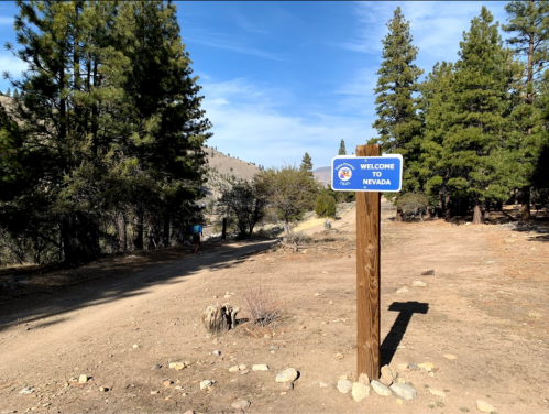 A wooden sign reading "Welcome to Nevada" stands on a dirt path surrounded by trees and mountains.
