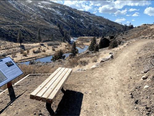 A wooden bench overlooks a winding river surrounded by hills and trees under a partly cloudy sky.