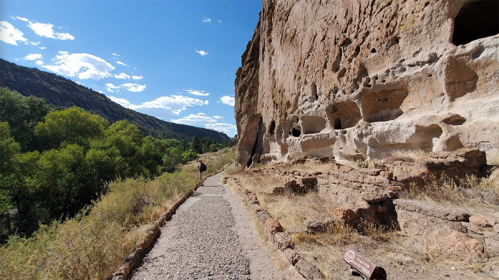 A scenic path alongside a rocky cliff with ancient cave dwellings and greenery under a blue sky with clouds.