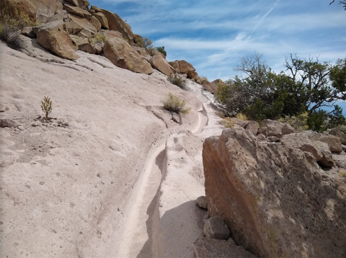 A sandy trail winds through rocky terrain under a blue sky, surrounded by sparse vegetation and boulders.