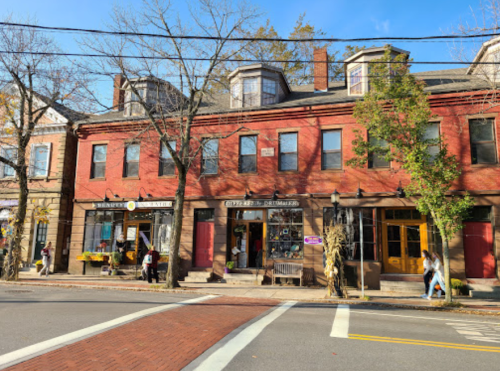 A charming street scene featuring red brick buildings with shops, trees, and pedestrians on a sunny day.