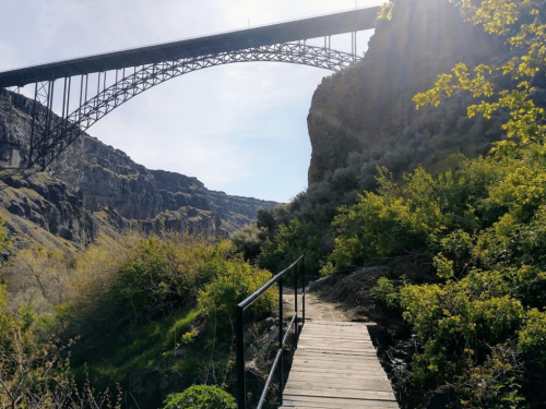 A wooden path leads through lush greenery beneath a large, arched bridge spanning a rocky canyon.