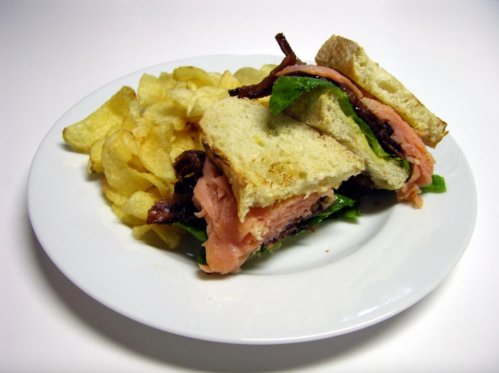 A sandwich with layers of ham, lettuce, and bacon, served with a side of potato chips on a white plate.