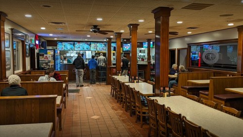 A cozy restaurant interior with wooden tables, patrons waiting in line, and multiple screens displaying menus.