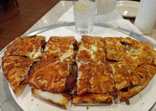 A large, cheesy pizza with various toppings, cut into slices, served on a metal tray with a glass of water.