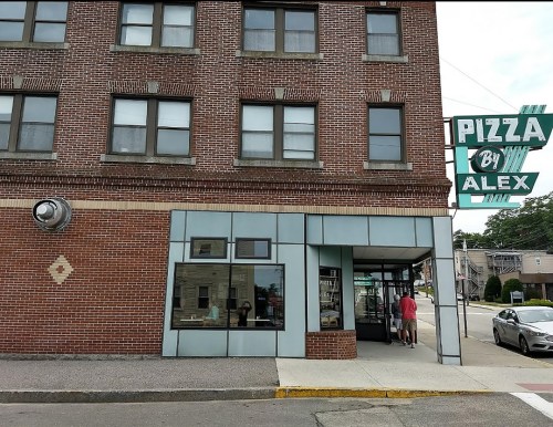 Exterior of a brick building with a sign reading "Pizza By Alex" and a person walking towards the entrance.