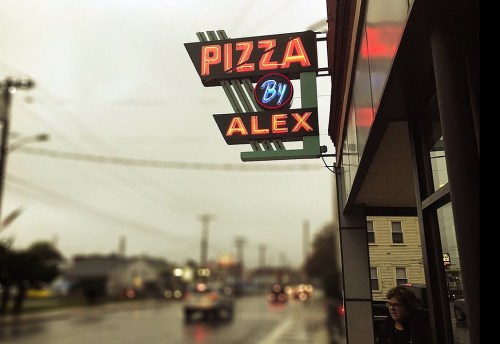 Neon sign for "Pizza by Alex" illuminated on a rainy street, with a person standing nearby.
