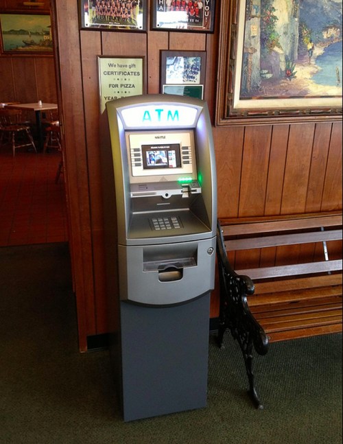 An ATM machine in a wooden-paneled interior, with a bench nearby and a sign about pizza gift certificates.