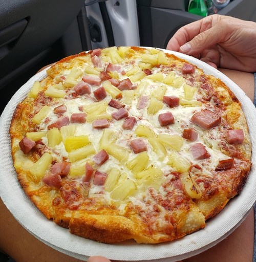 A close-up of a pizza topped with ham and pineapple, resting on a paper plate in a car.