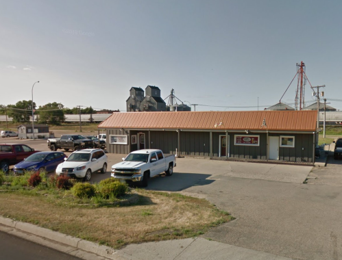 A small commercial building with a metal roof, surrounded by parked cars and industrial structures in the background.
