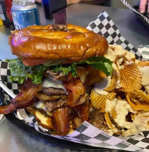 A towering burger with bacon, lettuce, and a pretzel bun, served with wavy chips drizzled in sauce.
