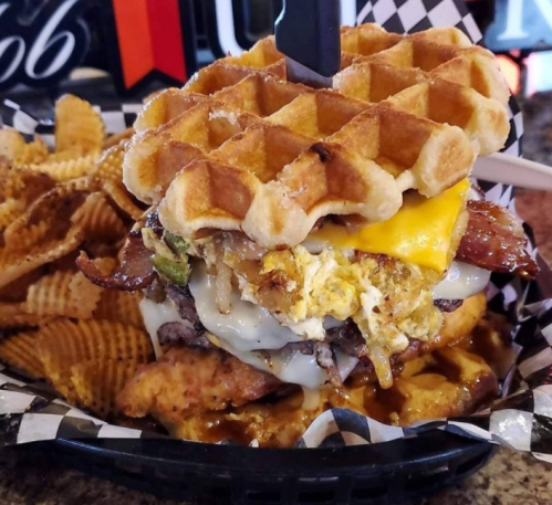 A towering waffle sandwich filled with bacon, cheese, eggs, and beef, served with crispy waffle fries on the side.