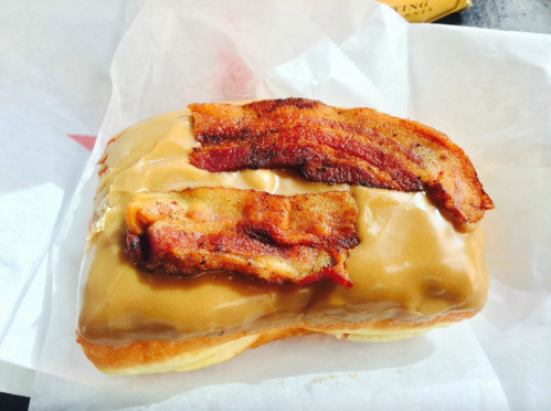 A glazed donut topped with crispy bacon strips on a white paper background.