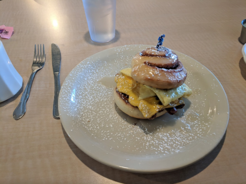 A plate with a stacked breakfast sandwich featuring a cinnamon roll, eggs, and cheese, dusted with powdered sugar.