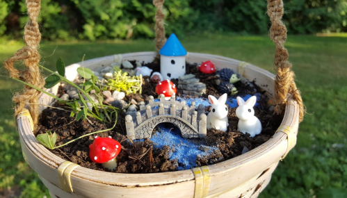 A whimsical miniature garden in a hanging pot, featuring tiny bunnies, mushrooms, a bridge, and a small house.