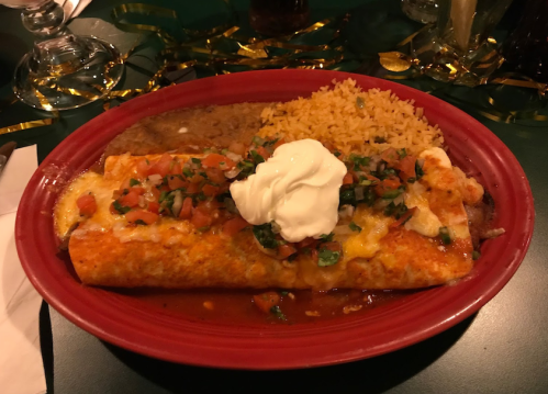 A red plate with two enchiladas topped with cheese, diced tomatoes, and sour cream, served with rice and refried beans.
