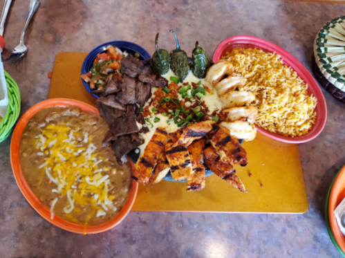 A colorful platter featuring grilled meats, shrimp, rice, beans, and fresh salsa, garnished with jalapeños and cheese.