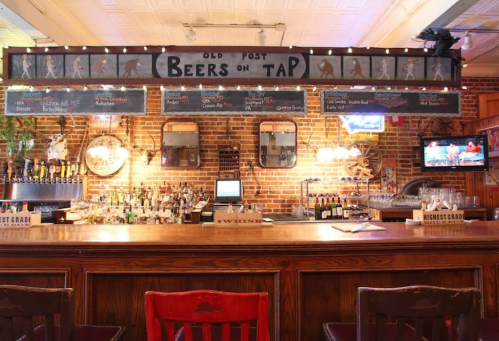A rustic bar with a wooden counter, beer taps, and a menu board above, featuring various beer options and a brick wall backdrop.