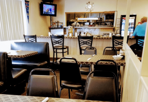 A cozy restaurant interior with black chairs and tables, a TV on the wall, and a view of the kitchen area.
