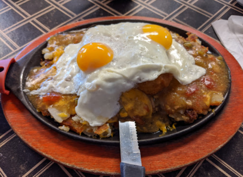 A sizzling plate of huevos rancheros topped with two fried eggs, surrounded by a mix of vegetables and cheese.
