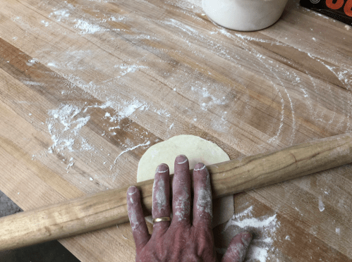 A hand rolls out dough on a wooden surface, surrounded by flour and kitchen tools.