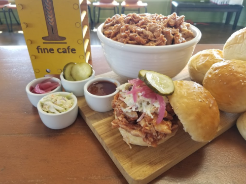 A plate featuring a pulled pork sandwich with pickles, coleslaw, and a bowl of shredded pork, alongside soft buns.
