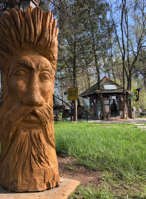 A carved wooden face stands in the foreground, with a rustic building and trees in the background.