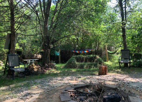 A serene outdoor space with a hammock, chairs, and a fire pit surrounded by trees and greenery.