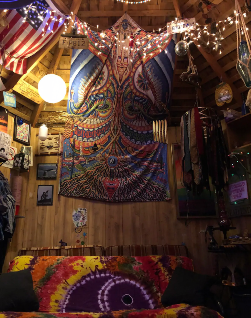 Colorful tapestry hangs on a wooden wall, surrounded by eclectic decor, string lights, and a tie-dye couch.