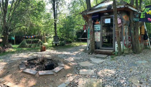 A rustic cabin in a wooded area, with a fire pit and stone path, surrounded by trees and colorful decorations.