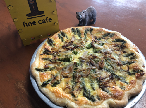 A freshly baked asparagus quiche on a plate, with a bear figurine and a sign for "fine cafe" in the background.