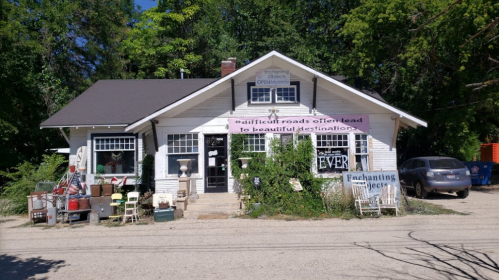 A charming white house with a sign, surrounded by greenery and various outdoor furniture, on a gravel road.