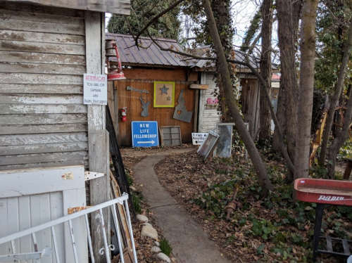 A rustic pathway leads to a colorful entrance with signs, surrounded by trees and old structures.