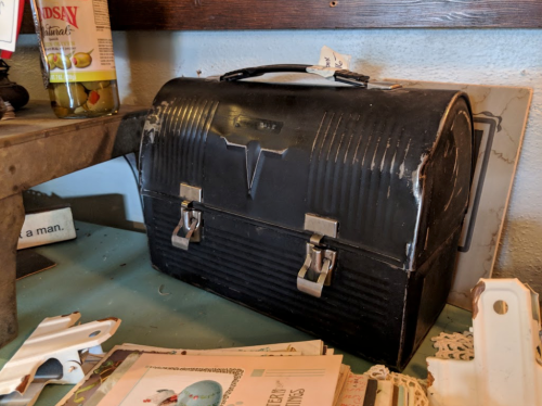 A vintage black toolbox with a curved top, metal latches, and a handle, resting on a table among various items.